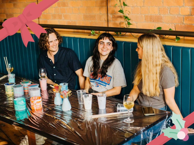 Three women smile and chat as they sit at a table containing resin art plant pots they've made at a craft class